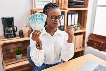 Sticker - African woman with braids working at the office holding brazilian reals pointing thumb up to the side smiling happy with open mouth