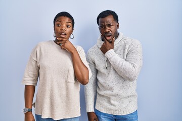 Poster - Young african american couple standing over blue background together looking fascinated with disbelief, surprise and amazed expression with hands on chin