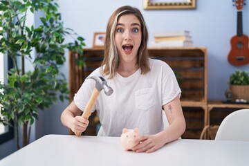 Poster - Blonde caucasian woman holding piggy bank and hammer celebrating crazy and amazed for success with open eyes screaming excited.