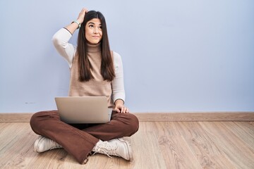 Canvas Print - Young brunette woman working using computer laptop sitting on the floor confuse and wondering about question. uncertain with doubt, thinking with hand on head. pensive concept.