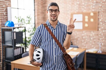 Canvas Print - Hispanic man with long hair working at the office holding bike helmet smiling with happy face winking at the camera doing victory sign with fingers. number two.