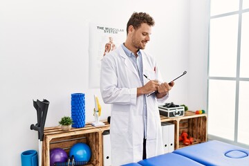 Sticker - Young hispanic man wearing physiotherapist uniform writing on clipboard at clinic