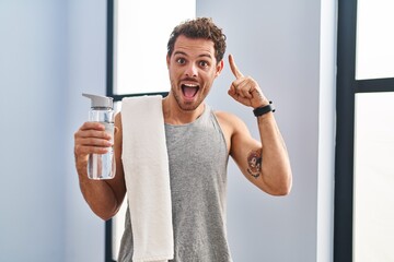 Sticker - Young hispanic man wearing sportswear drinking water smiling amazed and surprised and pointing up with fingers and raised arms.