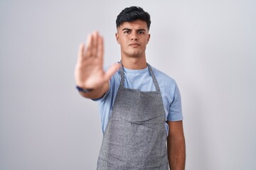 Sticker - Hispanic young man wearing apron over white background doing stop sing with palm of the hand. warning expression with negative and serious gesture on the face.