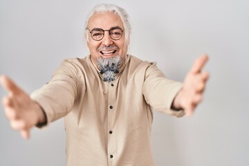 Wall Mural - Middle age man with grey hair standing over isolated background looking at the camera smiling with open arms for hug. cheerful expression embracing happiness.