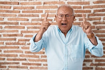 Canvas Print - Senior man with grey hair standing over bricks wall smiling amazed and surprised and pointing up with fingers and raised arms.