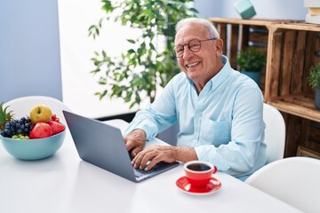 Wall Mural - Senior grey-haired man using laptop and drinking coffee sitting on table at home