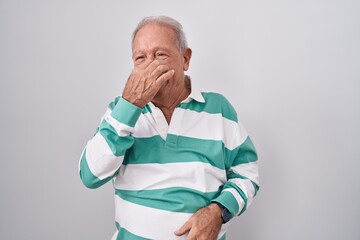 Poster - Senior man with grey hair standing over white background smelling something stinky and disgusting, intolerable smell, holding breath with fingers on nose. bad smell