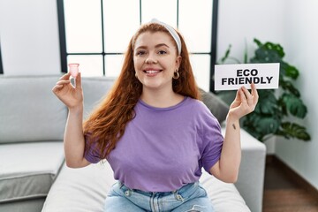 Wall Mural - Young redhead woman smiling confident holding menstrual cup and eco friendly banner at home