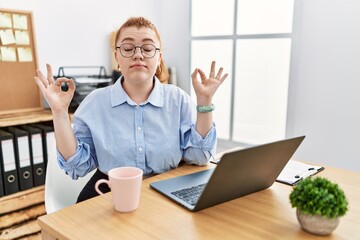 Canvas Print - Young redhead woman working at the office using computer laptop relaxed and smiling with eyes closed doing meditation gesture with fingers. yoga concept.