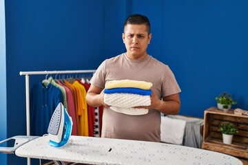 Poster - Hispanic young man holding folded laundry after ironing skeptic and nervous, frowning upset because of problem. negative person.