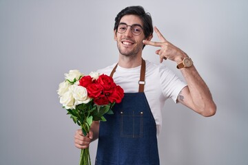 Poster - Young hispanic man holding bouquet of white and red roses doing peace symbol with fingers over face, smiling cheerful showing victory