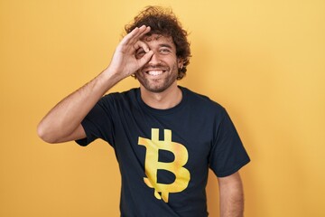 Canvas Print - Hispanic young man wearing bitcoin t shirt doing ok gesture with hand smiling, eye looking through fingers with happy face.