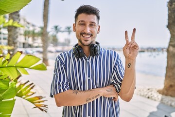 Canvas Print - Young handsome man listening to music using headphones outdoors smiling with happy face winking at the camera doing victory sign. number two.