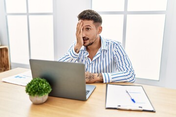 Wall Mural - Young handsome man with beard working at the office using computer laptop covering one eye with hand, confident smile on face and surprise emotion.