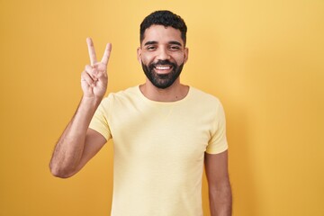 Poster - Hispanic man with beard standing over yellow background showing and pointing up with fingers number two while smiling confident and happy.