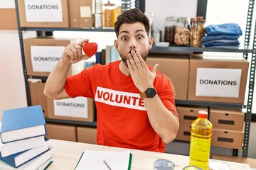 Canvas Print - Young man with beard at donations stand holding red heart covering mouth with hand, shocked and afraid for mistake. surprised expression