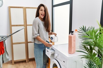 Poster - Young brunette woman putting detergent in washing machine celebrating crazy and amazed for success with open eyes screaming excited.