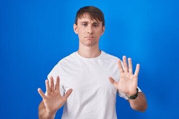 Poster - Caucasian blond man standing over blue background moving away hands palms showing refusal and denial with afraid and disgusting expression. stop and forbidden.