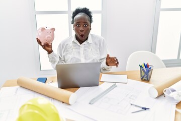 Wall Mural - Young african woman working at architecture studio holding piggy bank scared and amazed with open mouth for surprise, disbelief face
