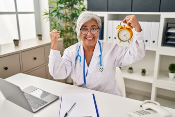 Poster - Middle age woman with grey hair wearing doctor uniform holding alarm clock screaming proud, celebrating victory and success very excited with raised arms