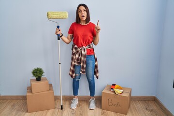 Sticker - Young hispanic woman painting home walls with paint roller amazed and surprised looking up and pointing with fingers and raised arms.