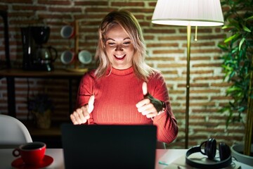 Canvas Print - Blonde woman using laptop at night at home approving doing positive gesture with hand, thumbs up smiling and happy for success. winner gesture.