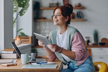 Wall Mural - Beautiful young woman using digital tablet and smiling while sitting at the kitchen counter at home