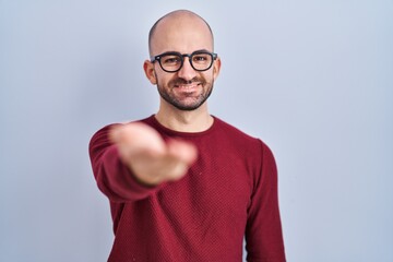 Sticker - Young bald man with beard standing over white background wearing glasses smiling cheerful offering palm hand giving assistance and acceptance.