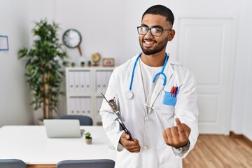 Sticker - Young indian man wearing doctor uniform and stethoscope beckoning come here gesture with hand inviting welcoming happy and smiling