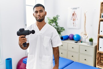 Wall Mural - Young indian physiotherapist holding therapy massage gun at wellness center making fish face with lips, crazy and comical gesture. funny expression.