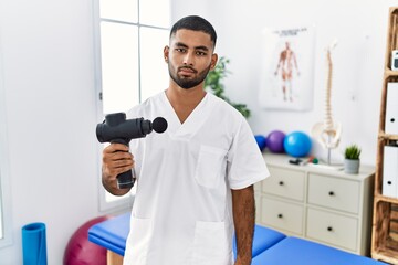 Wall Mural - Young indian physiotherapist holding therapy massage gun at wellness center relaxed with serious expression on face. simple and natural looking at the camera.