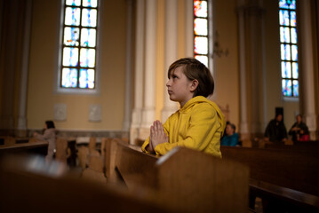 a little boy in a jacket prays in a dark catholic church at a children's mass