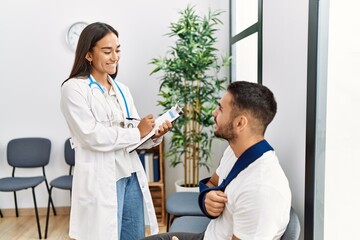 Poster - Youg latin injuried man wearing sling talking with the doctor at clinic waiting room.