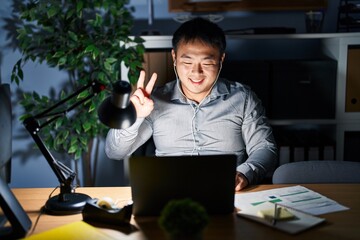 Canvas Print - Young chinese man working using computer laptop at night showing and pointing up with fingers number two while smiling confident and happy.