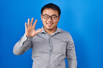 Sticker - Young chinese man standing over blue background showing and pointing up with fingers number five while smiling confident and happy.