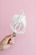 Close up of a hand balloon whisk with beaten egg white raw meringue for perfect peaks on pink background. High quality photo