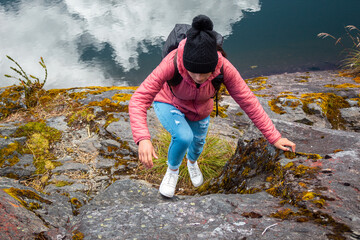 
turista con una chaqueta rosa escalando una roca junto a un lago tranquilo con nubes reflexionando sobre ella