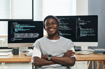 happy young successful it engineer in casualwear looking at you while sitting by workplace against c