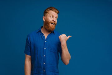 Portrait of adult handsome stylish redhead bearded surprised man in blue shirt with opened mouth,looking and pointing aside with thumb finger , while standing over isolated blue background