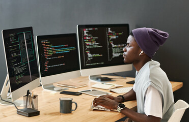 young serious it engineer in casualwear looking at coded data on computer screens while typing on ke