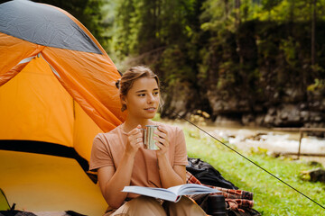 Wall Mural - White young woman reading book while resting by tent in green forest