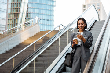 Wall Mural - Young woman using cellphone and holding coffee while going down escalator