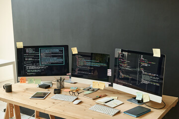 Three computer monitors with coded data and sticky notepapers on their screens standing on workplace of modern IT engineer in office