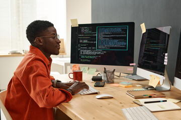 Young serious IT engineer in smart orange shirt looking at coded data on computer screens while typing on keyboard by workplace