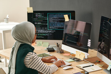 Young woman in hijab typing on computer keyboard while sitting by workplace in front of monitor in office and decoding data