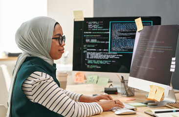 Young Muslim businesswoman in hijab looking at computer screen while typing and decoding data in front of monitor in office