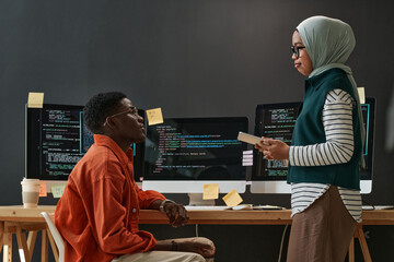 Two young confident multicultural interns in casualwear looking at each other during discussion of working points at meeting