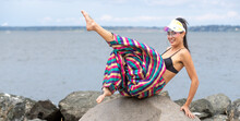 Asian Woman With Bare Feet Wearing Visor And Colorful Puffy Pants With A Bikini Top At A Waterfront Park On A Rock