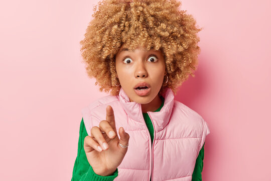 Photo of amazed curly haired young European woman keeps finger raised holds breath cannot believe own eyes wears green jumper and vest poses against pink background. People and reaction concept